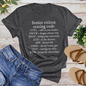 Op de afbeelding: Een donkergrijs T-shirt met witte tekst die "Senior citizen texting code" luidt en verschillende afkortingen en hun betekenissen opsomt. De afkortingen zijn: TTYL, FWTW, BYOT, ATD, BFF, OMSG, TOT, BTW.