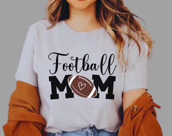May include: Light gray t-shirt with the words "Football Mom" in black script. The word "MOM" is in bold black letters, with a football graphic in the center. The football is brown with white stripes and a heart.
