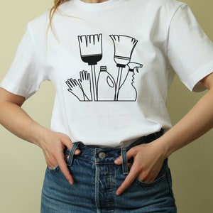May include: White t-shirt with a black graphic of cleaning supplies, including a broom, a spray bottle, gloves, and a bottle of cleaning solution.