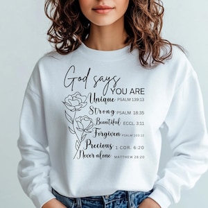 May include: White sweatshirt with black text that reads "God says you are Unique, Strong, Beautiful, Forgiven, Precious, Never alone" with bible verses listed below each word. The text is surrounded by a floral design.