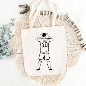 May include: White canvas tote bag with a black and white illustration of a soccer player with the number 10 and the name Messi on the back of the jersey.