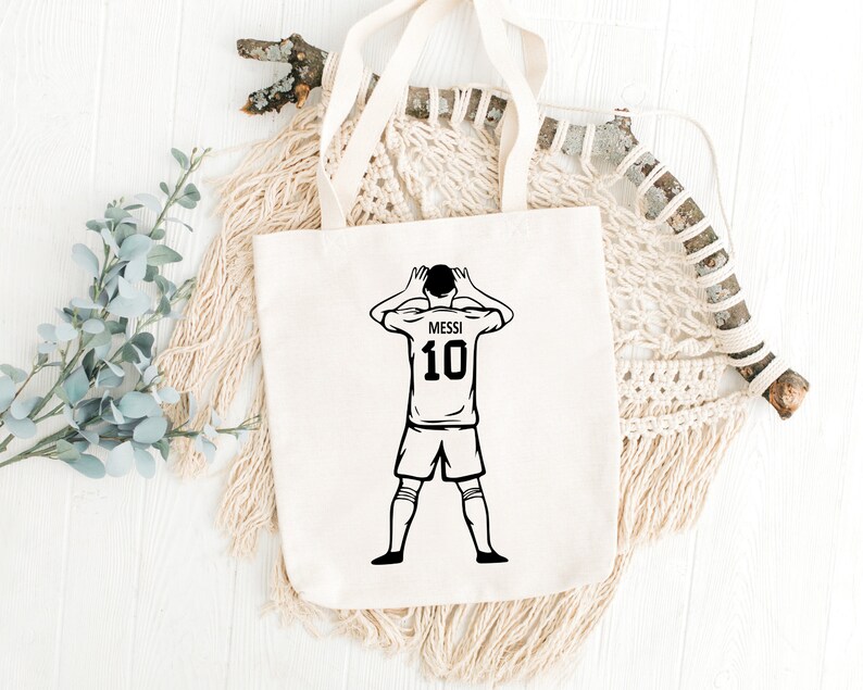 May include: White canvas tote bag with a black and white illustration of a soccer player with the number 10 and the name Messi on the back of the jersey.