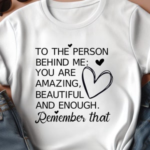 K&ouml;nnte beinhalten: Wei&szlig;es T-Shirt mit schwarzem Text, der lautet: "To the person behind me: You are amazing, beautiful and enough. Remember that". Der Text wird von kleinen Herzen begleitet.