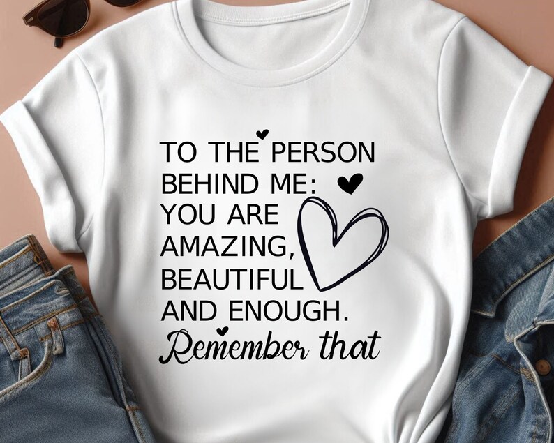 K&ouml;nnte beinhalten: Wei&szlig;es T-Shirt mit schwarzem Text, der lautet: "To the person behind me: You are amazing, beautiful and enough. Remember that". Der Text wird von kleinen Herzen begleitet.