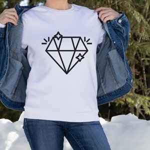 May include: A white sweatshirt featuring a black diamond graphic and two small stars. The sweatshirt is being held by a person wearing a denim jacket and blue jeans.