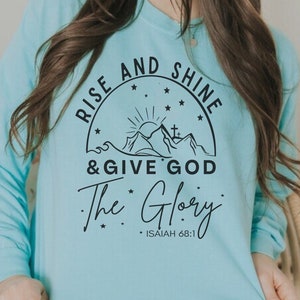 May include: A light blue sweatshirt with a black graphic design that says "Rise and Shine & Give God The Glory" with a mountain scene and stars. The text "Isaiah 68:1" is also included.