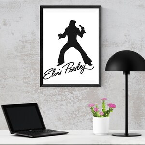 May include: Black and white silhouette of Elvis Presley singing with his signature on a white background in a black frame. A black lamp with a dome-shaped shade and a white planter with pink flowers are on a white surface.