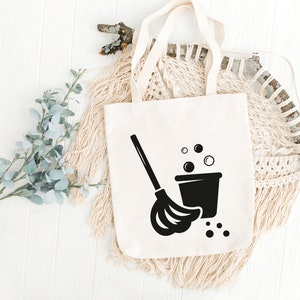 May include: A white canvas tote bag with a black silhouette of a mop and bucket with soap bubbles. The bag has a black handle.