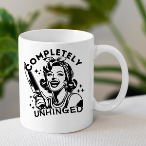 May include: White ceramic mug with a black and white illustration of a woman holding a knife. The text "COMPLETELY UNHINGED" is printed on the mug.