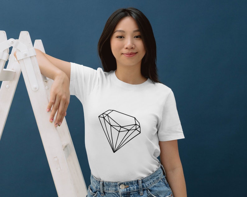 May include: A white t-shirt featuring a black geometric diamond design. The model is wearing the shirt and leaning against a white ladder.