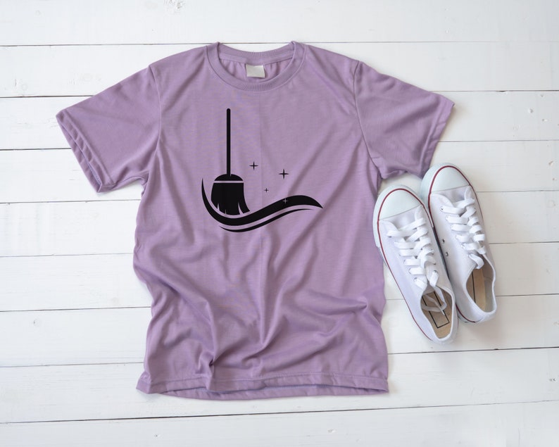 May include: A purple t-shirt with a black silhouette of a broom and stars on the front. The shirt is laid flat on a white wooden surface with a pair of white sneakers beside it.