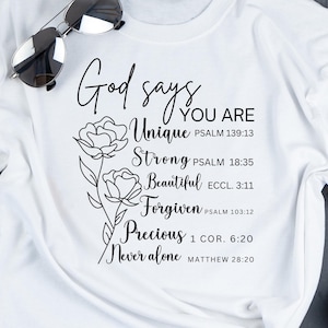May include: White t-shirt with black text that reads "God says you are Unique, Strong, Beautiful, Forgiven, Precious, Never alone" with bible verses listed below each word. The text is surrounded by a black line drawing of two flowers.