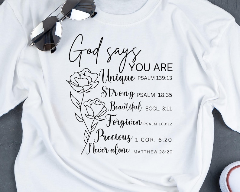 May include: White t-shirt with black text that reads "God says you are Unique, Strong, Beautiful, Forgiven, Precious, Never alone" with bible verses listed below each word. The text is surrounded by a black line drawing of two flowers.