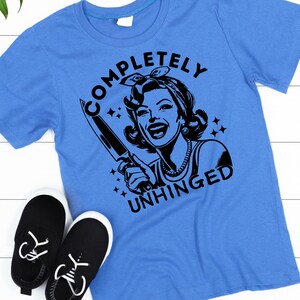 May include: A blue t-shirt with a black graphic of a woman holding a knife and the text "COMPLETELY UNHINGED".