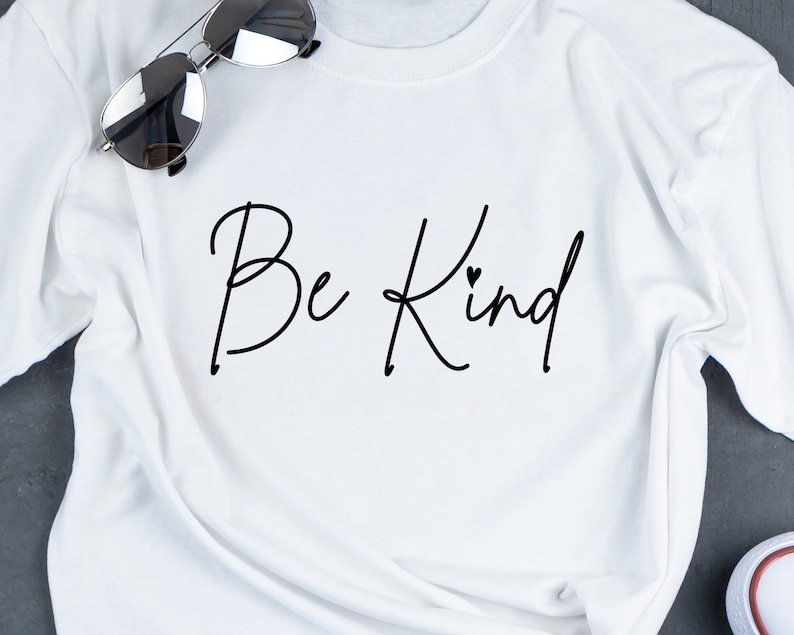 K&ouml;nnte beinhalten: Wei&szlig;es T-Shirt mit dem Text "Be Kind" in schwarzer Kursivschrift mit einem kleinen Herzen &uuml;ber dem "d".