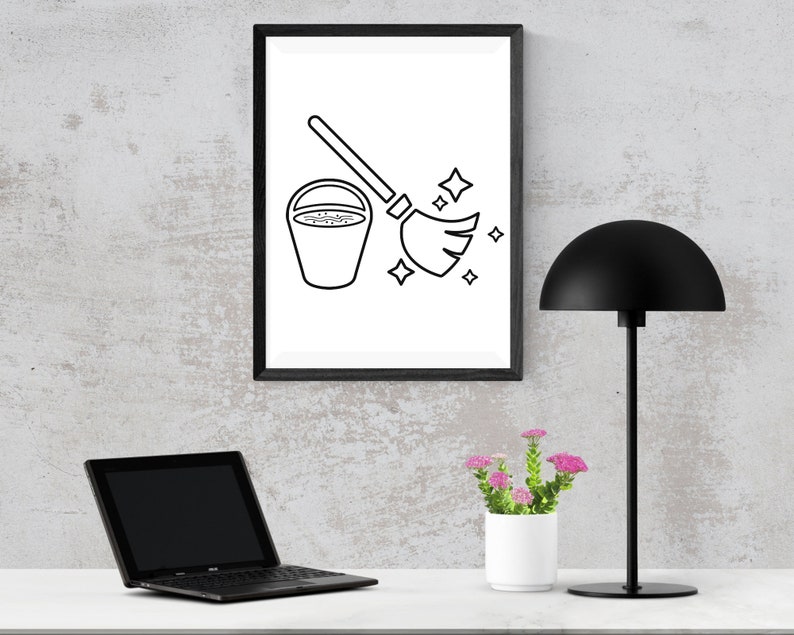 May include: A black and white line drawing of a bucket and broom with sparkling stars, framed in a black frame and hanging on a gray wall. A black lamp with a dome-shaped shade sits on a white surface next to a potted plant with pink flowers.