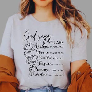 May include: White t-shirt with black text that reads "God says You Are Unique, Strong, Beautiful, Forgiven, Precious, Never Alone" with bible verses listed below each word. The text is surrounded by two stylized flower drawings.