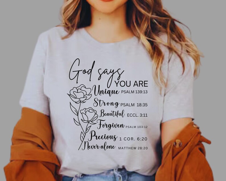 May include: White t-shirt with black text that reads "God says You Are Unique, Strong, Beautiful, Forgiven, Precious, Never Alone" with bible verses listed below each word. The text is surrounded by two stylized flower drawings.
