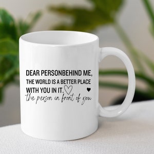 K&ouml;nnte beinhalten: Wei&szlig;e Keramik-Kaffeetasse mit schwarzem Text, der lautet: "Liebe Person hinter mir, die Welt ist ein besserer Ort mit dir drin. Die Person vor dir." Am Ende des Textes befinden sich zwei kleine schwarze Herzen.