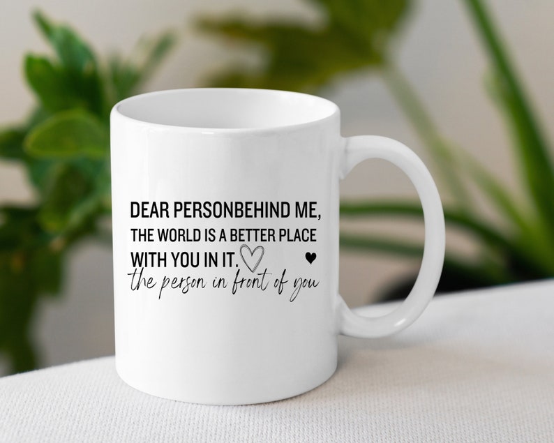 K&ouml;nnte beinhalten: Wei&szlig;e Keramik-Kaffeetasse mit schwarzem Text, der lautet: "Liebe Person hinter mir, die Welt ist ein besserer Ort mit dir drin. Die Person vor dir." Am Ende des Textes befinden sich zwei kleine schwarze Herzen.