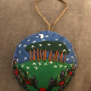 May include: A circular felt ornament with an embroidered landscape design. It depicts a blue sky with a crescent moon and stars, a green hill with trees, and floral embroidery. A jute twine loop is attached for hanging.