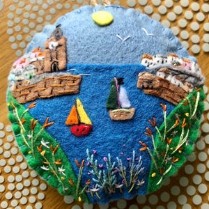 May include: A circular felt decoration showing a coastal scene. The blue sea has two sailboats and is bordered by green grass and embroidered flowers. Brown structures and buildings are on the left and right. A yellow sun and two birds are in the sky.