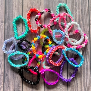 May include: A collection of colorful rubber band bracelets in various styles and patterns. The bracelets are made with different colors of rubber bands, including pink, blue, green, purple, and yellow. Some of the bracelets have beads or charms.