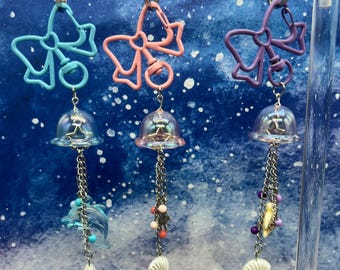 May include: Three iridescent glass bell charms with different colored bows and dangling charms. The charms are attached to silver keychains. The charms feature a blue dolphin, a pink starfish, and a purple seashell.