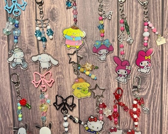 May include: A collection of 14 colorful keychains featuring various cartoon characters, including Hello Kitty, My Melody, and Pochacco. Each keychain is made with clear acrylic charms, beads, and metal rings. The keychains are displayed against a wooden background.