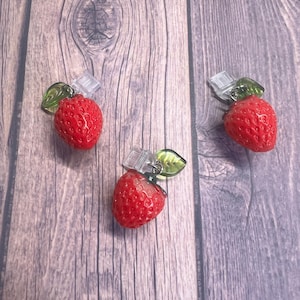 May include: Three red strawberry charms with green leaf accents and clear clasps. The charms are detailed with small bumps, resembling the texture of real strawberries. The background is a wooden surface.