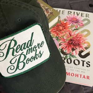 Puede incluir: Gorra de béisbol verde oscuro con un parche blanco que dice "Read more Books" en letras verdes. También se ve un libro con una portada floral y una camisa gris.