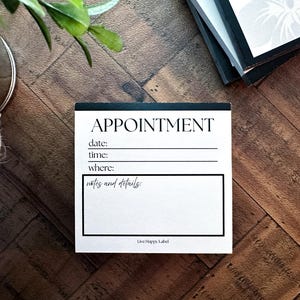 May include: A white appointment notepad with black text, including fields for date, time, and location. The word "APPOINTMENT" is at the top. The notepad is on a wooden surface.
