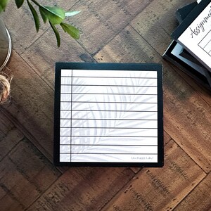May include: A black and white notepad with a palm leaf design and horizontal lines. The notepad is in a black box, with the text "Live Happy Labs" at the bottom. Other notepads are visible in the background.