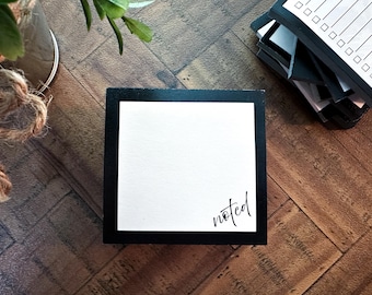 May include: A black and white notepad with the word "noted" in script. The notepad is on a wooden surface, with a stack of to-do list notepads in the background. The notepad is a square shape.