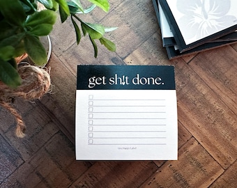 May include: A white notepad with the text "get sh!t done" in a black rectangle at the top. The notepad has a checklist format with lines and check boxes. The notepad is on a wooden surface.