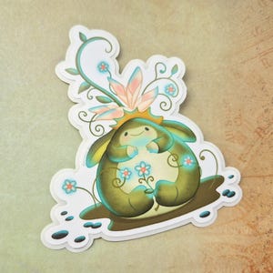 May include: A whimsical sticker featuring a cartoon creature with a smiling face, holding flowers. The creature is primarily olive green and cream, with blue and pink floral accents. The sticker has a white border and is set against a neutral background.