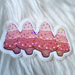 May include: A sticker featuring four stylized Christmas trees in shades of pink and red, with a gradient effect and glitter accents. The trees have a layered design with white dots and brown sprinkles.
