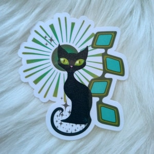 May include: A black cat with green eyes sits in front of a green and gold sunburst design. The cat has a sparkly tail and is surrounded by turquoise blue geometric shapes.