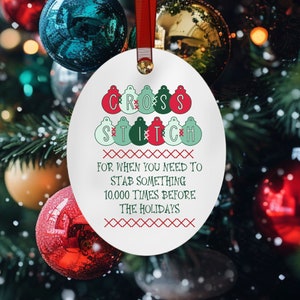 May include: Oval Christmas ornament with the words "CROSS STITCH" and the phrase "FOR WHEN YOU NEED TO STAB SOMETHING 10,000 TIMES BEFORE THE HOLIDAYS" in red, green, and white. The ornament hangs from a red ribbon.