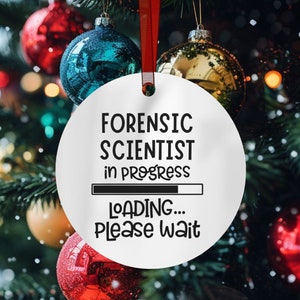 May include: White Christmas ornament with black text that reads "Forensic Scientist in Progress" with a progress bar and "Loading... Please wait". The ornament is hanging on a green Christmas tree with red and gold ornaments.
