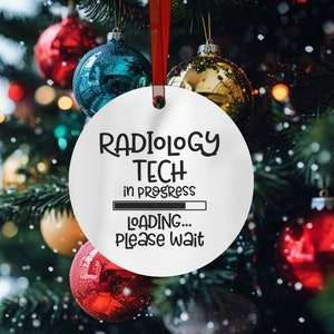 May include: White Christmas ornament with black text that reads "Radiology Tech in progress... Loading... Please wait". The ornament is hanging on a Christmas tree with red and gold ornaments.