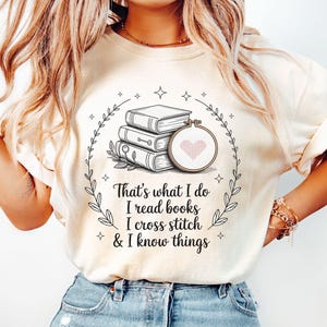 May include: Ivory-colored t-shirt featuring a graphic of stacked books and a cross-stitch hoop with a heart. The text reads, "That's what I do, I read books, I cross stitch & I know things."