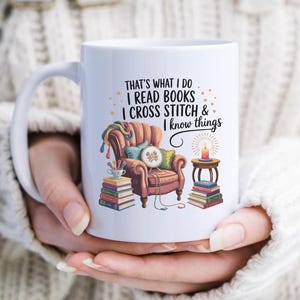May include: White ceramic mug featuring a cozy illustration of a reading chair, books, and a candle. The mug has the text "THAT'S WHAT I DO I READ BOOKS I CROSS STITCH & I know things". A perfect gift for book lovers and crafters.