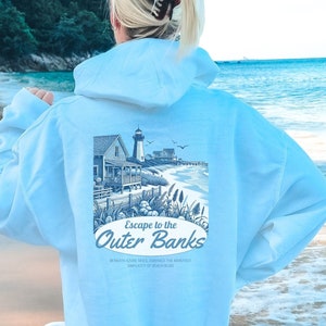 May include: Light blue hoodie with a graphic of a coastal scene, including a house, lighthouse, and the text "Escape to the Outer Banks." The hoodie is a light blue color and has a hood and long sleeves.