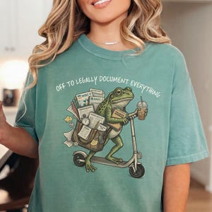 May include: Light green t-shirt with the text "OFF TO LEGALLY DOCUMENT EVERYTHING". The shirt features a cartoon frog riding a scooter, carrying a bag of documents and a coffee.