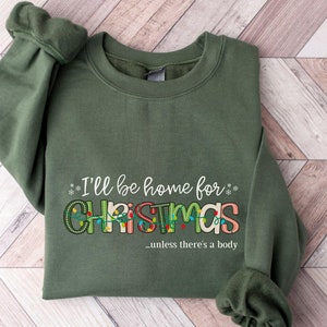 May include: Military green sweatshirt with the text "I'll be home for CHRISTMAS...unless there's a body" in colorful letters and Christmas lights. The sweatshirt is folded on a wooden surface.