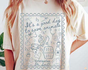 May include: A cream-colored t-shirt with a blue graphic of a postage stamp. The stamp features the text "It's a good day to save animals" and illustrations of a bone, a leash, a heart, a pet carrier, a bottle, a stethoscope, and a paw print. The text "Veterinarian Dr. Smith" is also included on the stamp.