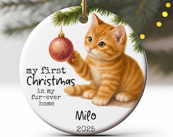 May include: A round Christmas ornament featuring a ginger kitten reaching for a red and gold ornament. Text on the ornament reads "my first Christmas in my fur-ever home" and includes the name "Milo" and the year "2025".