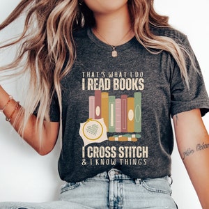 May include: A dark grey heather t-shirt with the text "That's what I do I read books I cross stitch & I know things" in white. The text is surrounded by a stack of colorful books and a cross stitch hoop.
