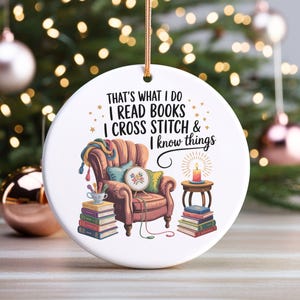 May include: A round white ceramic ornament featuring a graphic of a brown armchair, stacks of books, a lit candle on a side table, and a cross stitch hoop. The text reads: THAT'S WHAT I DO I READ BOOKS I CROSS STITCH & I know things.